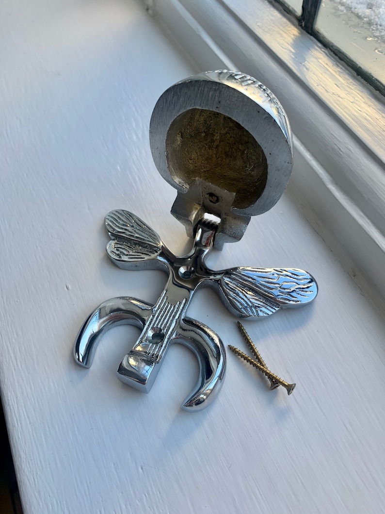 May include: A silver door knocker shaped like a butterfly with two wings. The knocker is open and shows the inside of the knocker, which is hollow. Two screws are included.