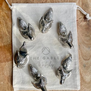 May include: Six silver fox head bottle stoppers on a white fabric background. The stoppers are all facing forward and have a detailed design. The text "The Gables Workshop" is printed on the fabric.