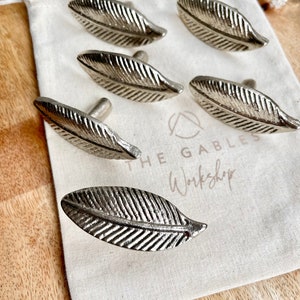 May include: Six silver leaf-shaped cabinet knobs in a white fabric bag. The bag has the text "The Gables Workshop" printed on it.