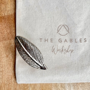 May include: A silver leaf-shaped door handle with a textured surface. The handle is on a white canvas bag with the text "THE GABLES Workshop" printed on it.