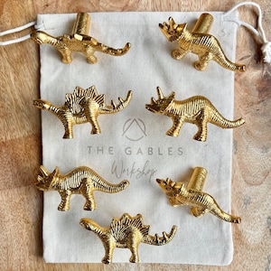 May include: Six gold-coloured dinosaur-shaped cabinet knobs. The knobs are presented on a canvas bag with the text "THE GABLES Workshop". The dinosaurs include a Stegosaurus, Triceratops, and other species.
