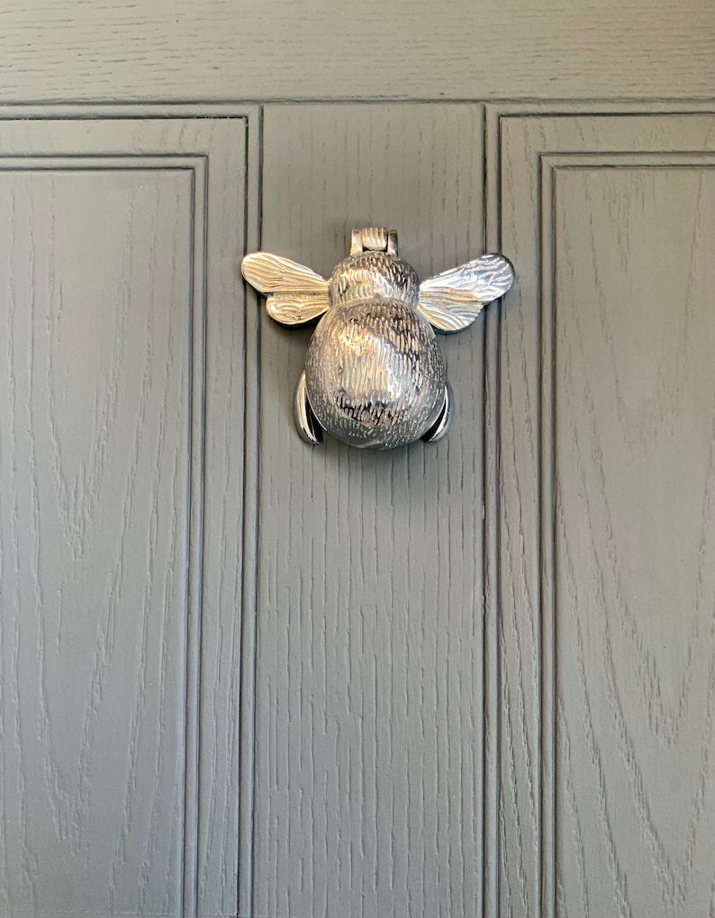 May include: A silver bee-shaped door knocker on a gray wooden door.