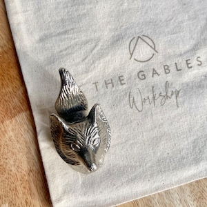 May include: A silver fox head bottle stopper with a detailed design. The stopper is sitting on a white fabric bag with the text "THE GABLES Workshop" printed on it.