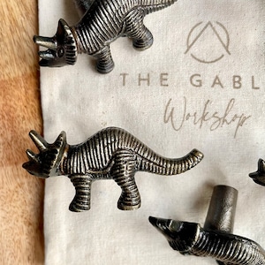 May include: A set of four antique bronze dinosaur-shaped cabinet knobs. The knobs are in the shape of a Triceratops dinosaur. The knobs are displayed on a white fabric surface with the text "THE GABL Workshop".