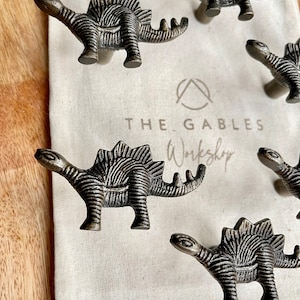 May include: Six antique silver dinosaur-shaped drawer pulls on a white canvas bag with the text "THE GABLES Workshop" printed on it.