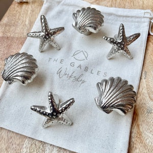 May include: Six silver cabinet knobs shaped as starfish and seashells, presented on a white drawstring bag. The bag has the text "THE GABLES Workshop". The background is a light-coloured wooden surface.