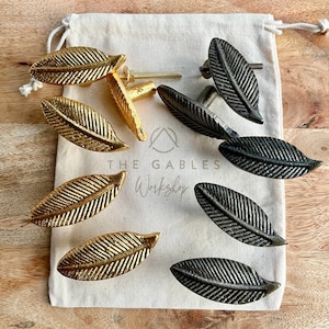 May include: A set of eight leaf-shaped cabinet knobs in two finishes: gold and black. The knobs are arranged in a white fabric bag with the text "THE GABLES Workshop" printed on it.