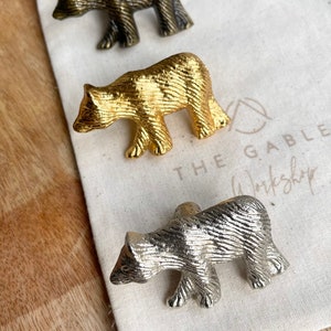 May include: Three bear-shaped cabinet knobs in different finishes: antique brass, gold, and silver. The knobs are displayed on a white fabric surface with the text "The Gable Workshop" printed on it.