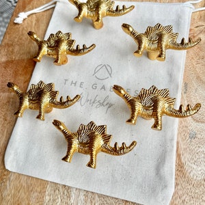 May include: Five gold dinosaur-shaped cabinet knobs on a white fabric surface. The knobs have a textured, scaly design and are perfect for adding a whimsical touch to any room.