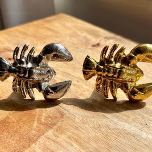 May include: Two decorative lobster figurines. One is silver, the other is gold. Both have detailed claws, segmented bodies, and antennae. They are displayed on a wooden surface.