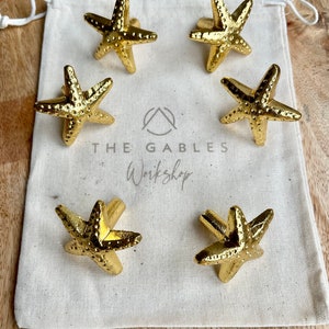 May include: Six gold-toned starfish drawer pulls in a white fabric pouch. The pouch has the text "The Gables Workshop" printed on it.