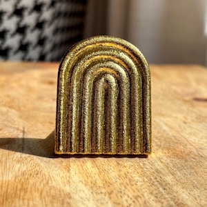 May include: A gold-coloured, arched decorative object with a textured surface. The object has a series of parallel, vertical lines that create a ribbed effect. The item is on a wooden surface.