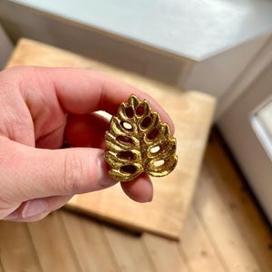 May include: A gold-coloured leaf-shaped decorative object. The leaf has a detailed, openwork design with multiple oval-shaped cutouts. The object is held in a hand, and the background is a wooden surface.