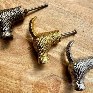 May include: Three dog-shaped cabinet pulls in gold, silver, and bronze finishes. Each pull has a textured body and a curved tail, with a threaded bolt for installation. The pulls are arranged on a wooden surface.