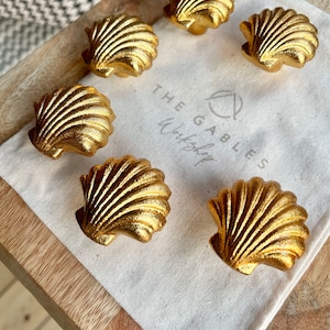 May include: Six gold seashell cabinet knobs displayed on a white cloth bag. The knobs are made of metal and have a detailed, textured design. The bag has the text "THE GABLES Workshop" printed on it.