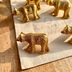 May include: A set of six gold bear-shaped drawer pulls. The pulls are made of metal and have a textured finish. The pulls are arranged on a white fabric surface with the text "The Gables Workshop" visible in the background.