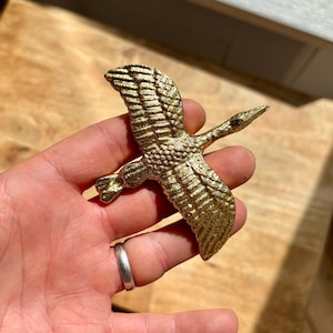 May include: A gold-coloured metal bird figurine in flight. The bird has detailed wing and feather textures, with a long neck and beak. The figurine is held against a wooden surface.