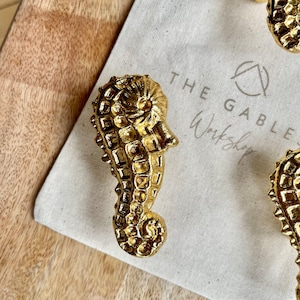 May include: Gold seahorse-shaped cabinet pulls. The seahorse hardware is detailed with textured surfaces and a curved tail. The pulls are displayed on a neutral canvas bag with the text "THE GABLE Workshop". The background is a light wood surface.