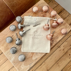 May include: A collection of 15 silver and copper cabinet knobs with a textured surface. The knobs are arranged on a wooden surface with a white fabric bag labelled "The Gables Workshop".