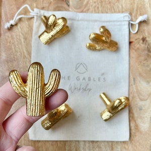 May include: Gold-coloured cactus-shaped cabinet knobs and pulls. The knobs are in various sizes and shapes, with a textured surface. The knobs are displayed with a small drawstring bag that has the text "THE GABLES Workshop".