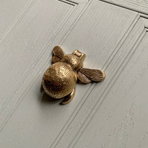 May include: A gold-toned bee-shaped door knocker with a textured body and detailed wings.