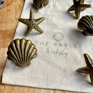 May include: A set of six gold-toned metal drawer pulls in the shape of seashells and starfish. The pulls are displayed on a white fabric bag with the text "THE GABLES Workshop" printed on it.