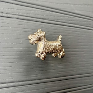 May include: A gold-coloured cabinet knob shaped like a Scottish Terrier dog. The dog is shown standing with detailed fur texture. The knob is mounted on a grey wooden surface.