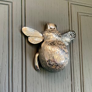 May include: A silver bee-shaped door knocker with detailed wings and a textured body.