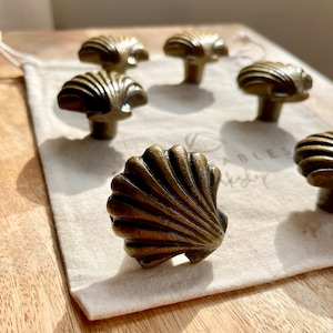 May include: Six antique gold-toned seashell cabinet knobs with a detailed design. The knobs are arranged on a white fabric surface.