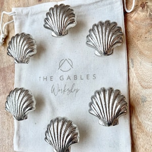 May include: Six silver seashell cabinet knobs displayed on a cream drawstring bag. The bag features the text "THE GABLES Workshop". The knobs are metallic and have a detailed, textured design. The background is a wooden surface.