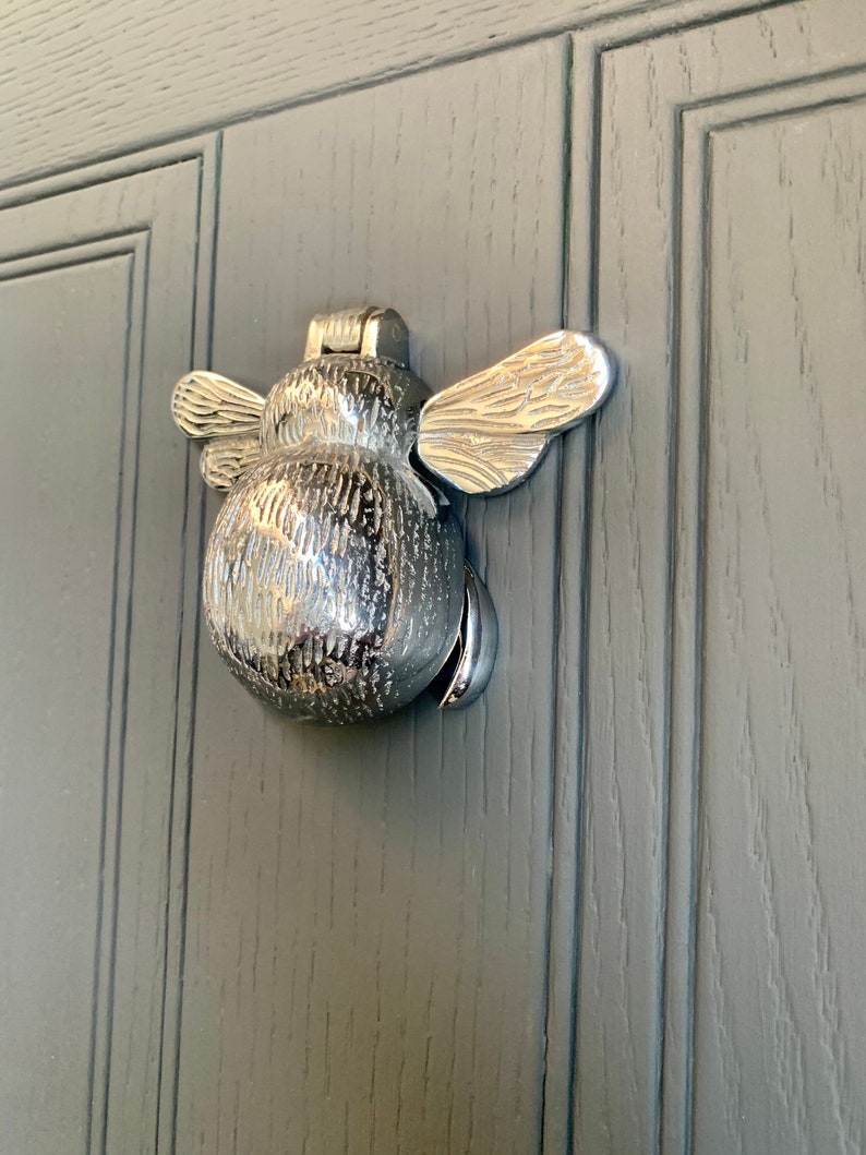 May include: A silver bee-shaped door knocker with textured wings and body. The knocker is mounted on a gray wooden door.