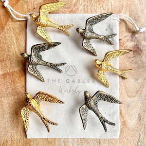 May include: Six gold and silver bird-shaped drawer pulls in a white fabric bag. The bag has the text "THE GABLES Workshop" printed on it.