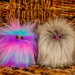 May include: Two fluffy, fuzzy, plush toys with large, round, black eyes and pink eyelids. The toy on the left is pink, blue, and purple. The toy on the right is gray.