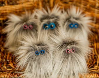 May include: Five fluffy, gray plush creatures with large, colorful eyes. The creatures have a shaggy, fur-like texture and are arranged in a wicker basket. The eyes are either pink or blue, adding a playful touch to the whimsical design.