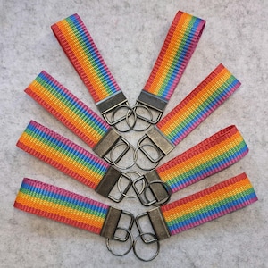 Könnte beinhalten: Eine Sammlung von sechs Schlüsselanhängern aus Stoff mit einem lebendigen Regenbogen-Streifenmuster. Jedes Band besteht aus gewebtem Material und ist an einem dunklen, rechteckigen Metallverschluss mit einem runden Schlüsselring befestigt.