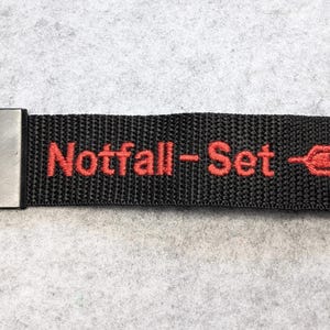 Könnte beinhalten: Schwarzer Schlüsselanhänger mit dem rot gestickten Text "Notfall-Set" und einem Spritzen-Symbol. Der Riemen hat einen Metallverschluss und einen schwarzen Metallring zum Befestigen von Schlüsseln.