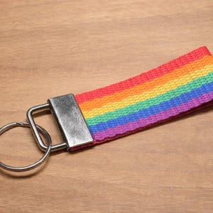 Könnte beinhalten: Ein Schlüsselanhänger mit einem flachen, gewebten Stoffband mit horizontalen Streifen in Rot, Orange, Gelb, Grün, Blau und Violett. Das Band ist an einer rechteckigen silberfarbenen Metallklemme und einem runden Schlüsselring befestigt.