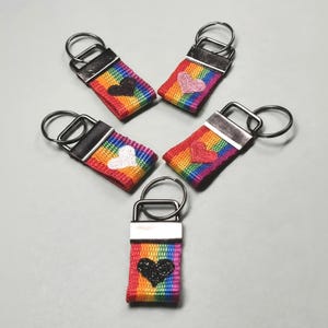 Könnte beinhalten: Fünf Schlüsselanhänger mit regenbogenfarbenen Stoffbändern, silberfarbenen Metallbeschlägen und runden Ringen. Jedes Band ist mit einem einzelnen gestickten Herz in verschiedenen Farben versehen, darunter Schwarz, Rosa, Weiß, Rot und Glitzerschwarz.