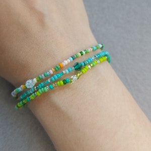 Könnte beinhalten: Ein Set aus drei Perlenarmbändern in Grün-, Weiß- und Rosatönen. Die Armbänder sind übereinander am Handgelenk einer Person gelegt.