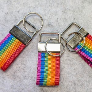 Könnte beinhalten: Drei Schlüsselanhänger mit einem regenbogenfarbenen Stoffband aus gewebtem Nylon. Jeder Schlüsselanhänger verfügt über eine rechteckige Metallklemme in Silberoptik und einen runden Spaltring zur Befestigung von Schlüsseln. Die Streifen sind rot, orange, gelb, grün, blau und violett.