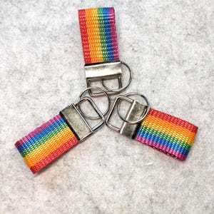 Könnte beinhalten: Drei Schlüsselanhänger aus regenbogenfarbenem, gewebtem Nylongewebe. Jeder Anhänger verfügt über eine silberfarbene Metallklemme an einem Ende, die die Stoffschlaufe sichert, sowie einen runden, silberfarbenen Metallring für Schlüssel.
