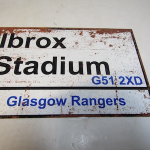 May include: A rectangular metal sign with a distressed, aged appearance. The sign features the words "Ibrox Stadium" in black, with "Glasgow Rangers" in blue. The sign also includes the postcode "G51 2XD". The edges show rust.