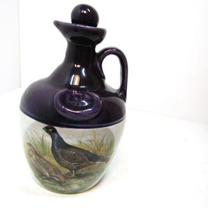 Può includere: Una caraffa in ceramica viola con un designo bianco e marrone che presenta un'illustrazione di un uccello. La caraffa ha un tappo di sughero e un manico.