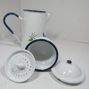 Op de afbeelding: Een witte geëmailleerde koffiepot met een blauwe rand en handvat, voorzien van een geschilderde eend en riet. De set bevat een bijpassende witte geëmailleerde zeefinzet met handvat en een los wit geëmailleerd deksel met een blauwe knop.