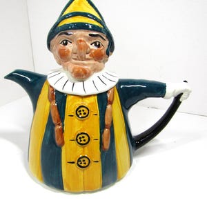 May include: A whimsical ceramic teapot in the shape of a jester. The teapot features a yellow and teal striped body, a jester's face on top, and a black handle. The jester wears a yellow and teal hat and a white ruffled collar.