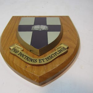 Op de afbeelding: Houten schild met een wapen met een blauw en zilver kruisontwerp en een open boek. Een gouden banner hieronder luidt "PRO DOCTRINIS ET DISCIPLINIS".