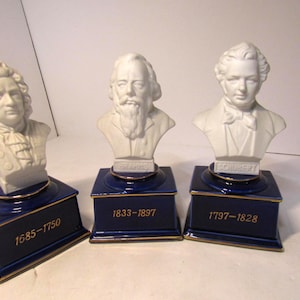 May include: Three white porcelain busts of composers, mounted on blue pedestals with gold trim. Each pedestal displays the composer's name and birth/death years: Bach, Brahms, and Schubert. A decorative collectible.