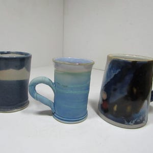 May include: Three ceramic mugs with distinct glazes. The left mug is two-toned blue and white. The center mug features a light blue and white gradient. The right mug has a dark blue and black mottled finish with small circular accents.