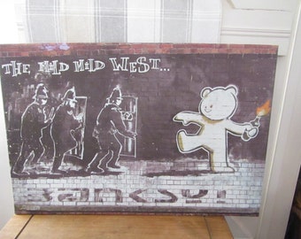 May include: A white bear with a Molotov cocktail runs away from three police officers in front of a brick wall. The text "THE MILD WILD WEST..." is above the bear and the text "BANKSY" is below the bear.
