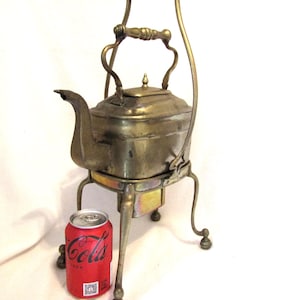 Puede incluir: Tetera antigua de latón con un soporte y un asa. La tetera está sobre un soporte con tres patas. Una lata de Coca-Cola está en primer plano para la escala.